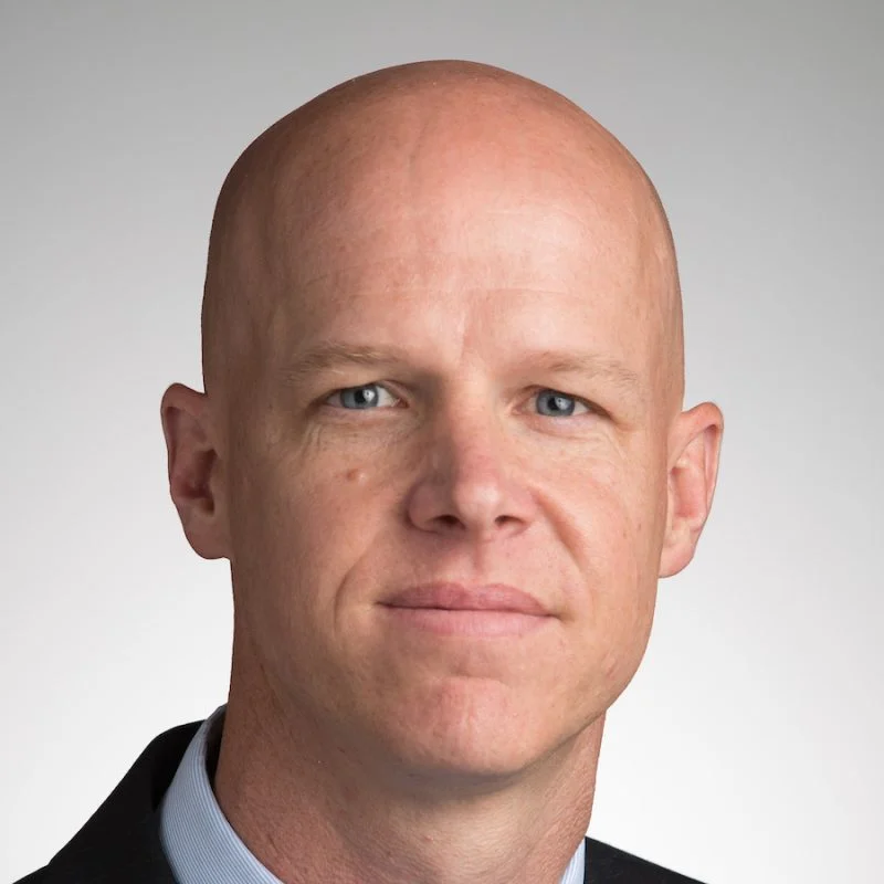 A bald man in a suit and collared shirt, resembling Matthew Puffer, looks at the camera with a neutral expression.