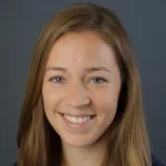 Smiling woman with straight brown hair and blue eyes against a plain dark background, Reva Johnson, Ph.D. appears confident and approachable.
