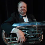 Richard Watson, dressed in a tuxedo, holds a large tuba and smiles against a dark background.