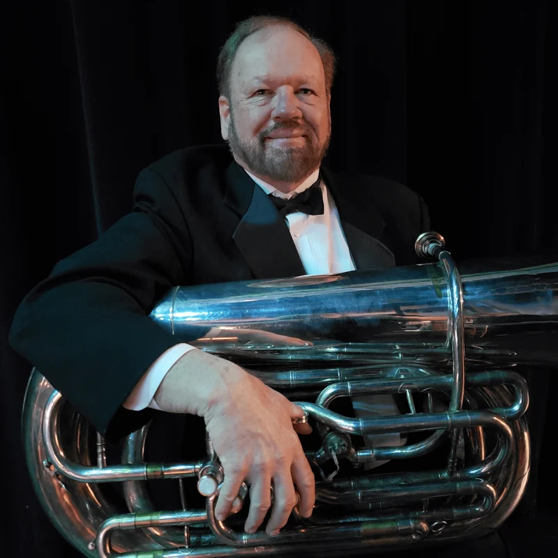 Richard Watson, dressed in a tuxedo, holds a large tuba and smiles against a dark background.