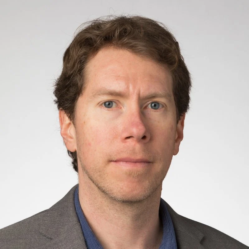 Giosuè Alagna, a man with light skin and wavy brown hair, wears a gray blazer and blue shirt against a plain background.