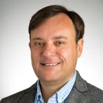 Carlos Miguel-Pueyo, a smiling man in a gray blazer and blue checkered shirt, stands against a plain, light background.