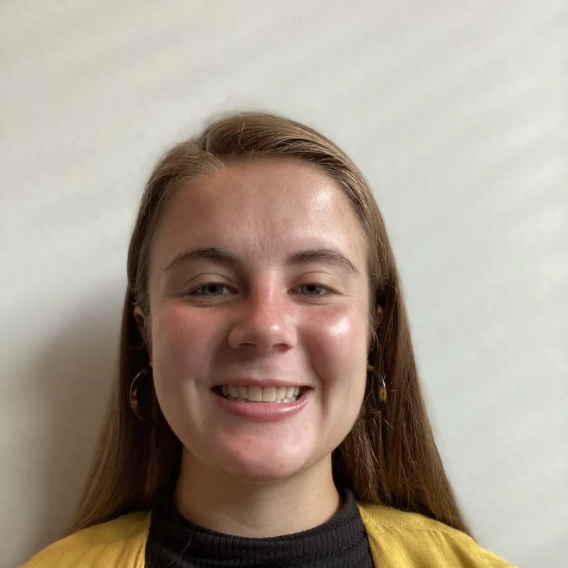 Young woman with long brown hair smiling at the camera, wearing a yellow cardigan and black top—Samantha Burgett.