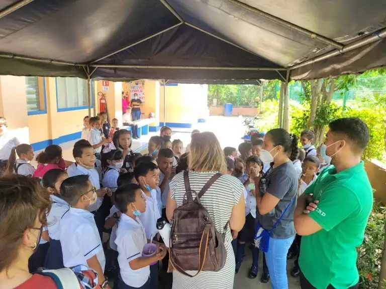 A group of school children and adults gather under a canopy outdoors, some wearing face masks as they watch WAVES of excitement ripple through the crowd.