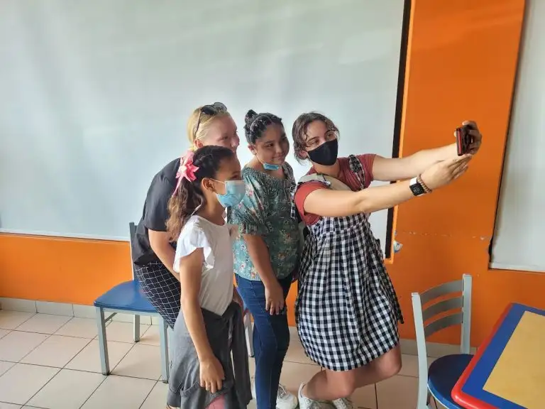 Four people wearing masks pose together for a selfie in a brightly colored room with orange and white walls, striking playful WAVES for the camera.