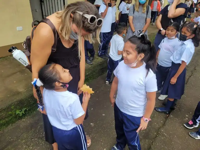 A woman waves as she talks to two young girls in school uniforms outside, with other children and adults in the background.