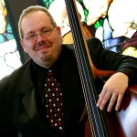 Phillip Serna, a smiling man in a suit, holds an upright bass with vibrant stained glass art in the background.