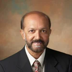 Shirvel Stanislaus, dressed in a suit and tie, smiles in a formal portrait with a neutral brown background.