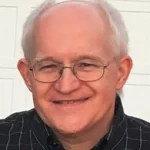 Smiling older man with glasses and short gray hair, wearing a black checkered shirt, stands in front of a white wall. This is Stan Zygmunt.