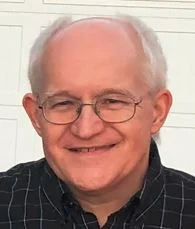 Smiling older man with glasses and short gray hair, wearing a black checkered shirt, stands in front of a white wall. This is Stan Zygmunt.