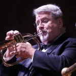 Charles Steck, dressed in a suit, plays a brass instrument—possibly a flugelhorn—captivating the audience during his performance.