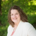 Smiling woman with curly brown hair in a white sweater, Stephanie Harris stands outdoors with greenery in the background.