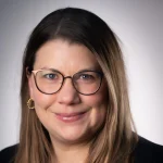 Smiling woman with long brown hair, glasses, and gold hoop earrings, in front of a neutral background—Theresa Carlson Carroll, OTD in occupational therapy.