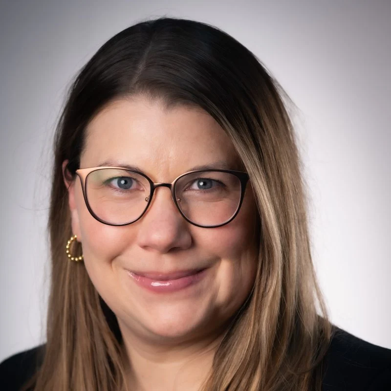 Smiling woman with long brown hair, glasses, and gold hoop earrings, in front of a neutral background—Theresa Carlson Carroll, OTD in occupational therapy.