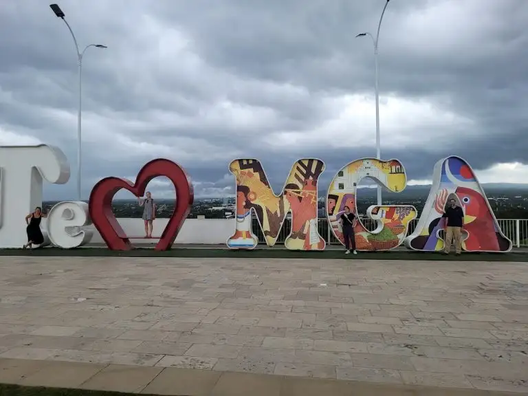 People pose in large colorful "Te Amo MGA" letters with a heart, as WAVES of clouds drift across the sky above the vibrant plaza.