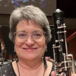 Smiling woman with short gray hair, possibly Barbara Timmerman, holds a woodwind instrument—perhaps an oboe—in a concert setting.