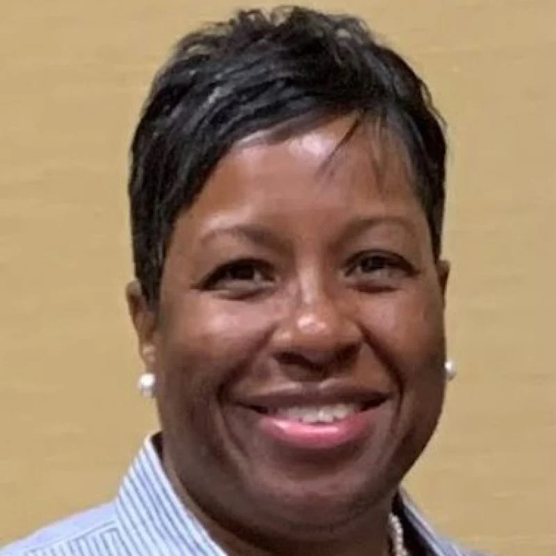 Victoria Baldwin, a woman with short dark hair, smiles warmly in a striped shirt and pearl earrings, posed against a beige background.