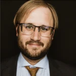 Robin Crigler, a man with glasses, a beard, and a suit, smiles against a dark background.