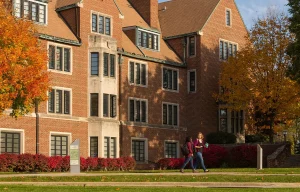 Two people walk past a large brick campus building surrounded by autumn trees and bushes.