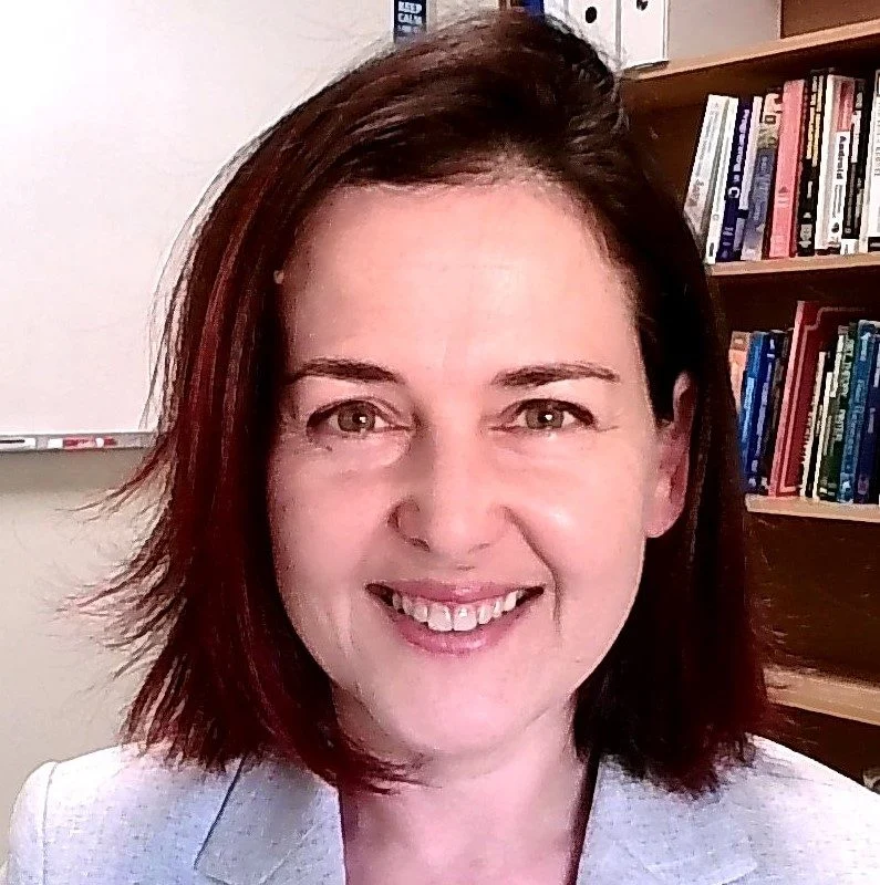 Aysegul Yayimli, a woman with shoulder-length brown hair, smiles while standing in front of bookshelves and a whiteboard.