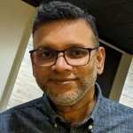 PhD holder Bharath Ganesh Babu, a man with glasses and a salt-and-pepper beard, smiles warmly while wearing a gray button-up shirt indoors.