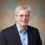 Edward Byrne, a man with gray hair and glasses, smiles warmly in a dark blazer and blue shirt, standing before a neutral backdrop.