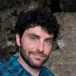Jarron Bowman, a man with curly dark hair and a beard, wearing a blue plaid shirt, stands in front of a rocky background.