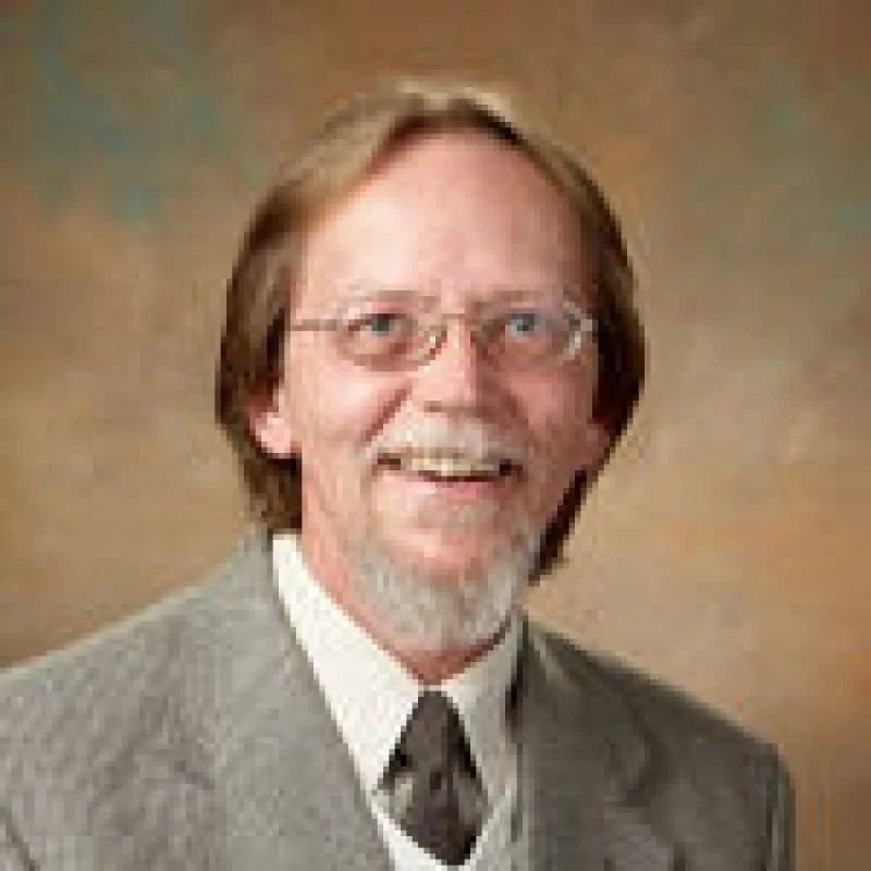 Smiling man with glasses, a beard, and mustache, wearing a gray suit and tie, poses against a brown background—Daniel Arkkelin, Ph.D.