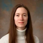 Melissa Desjarlais, a woman with long brown hair, wears a white turtleneck as she poses in front of a brown and blue background.