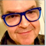 Smiling older man, Stephen John, with blue glasses, short gray hair, and a plaid shirt, facing the camera.