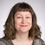Smiling woman with wavy brown hair and bangs, wearing a patterned top, against a light background.