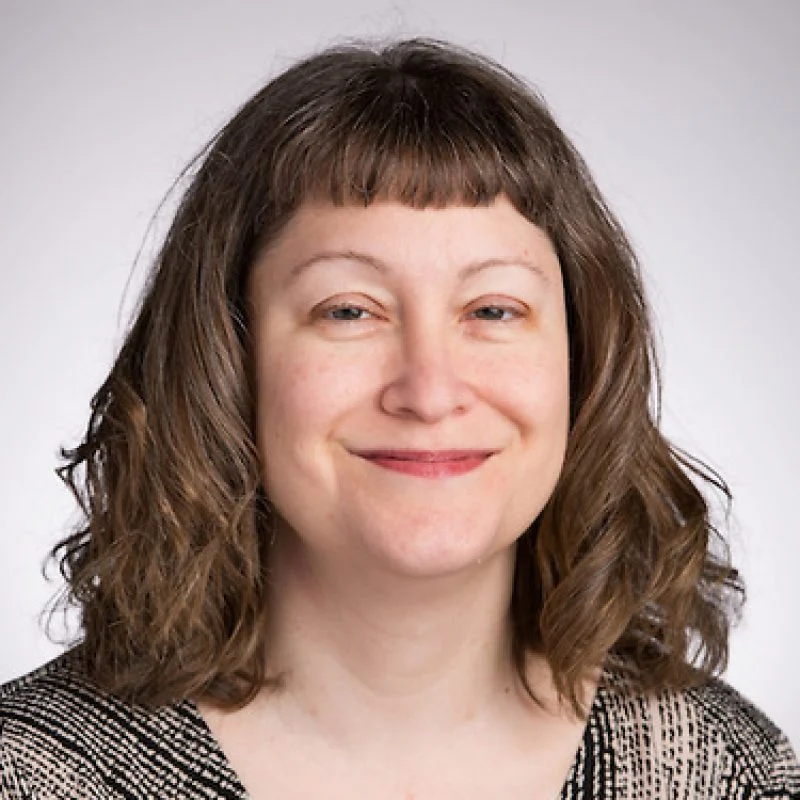 Smiling woman with wavy brown hair and bangs, wearing a patterned top, against a light background.