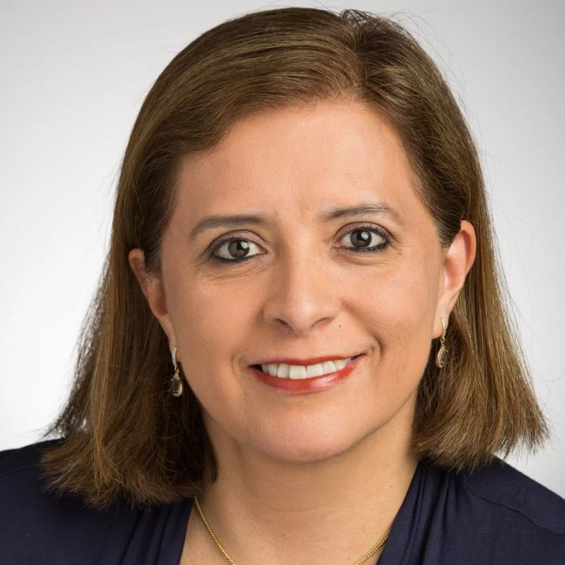 Smiling woman, Nelly Zamora-Breckenridge, with straight brown hair, wearing a dark top and gold jewelry, against a light background.