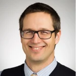 Christopher Iceman, a smiling man with short brown hair, glasses, a collared shirt, tie, and dark sweater vest, stands against a light background.
