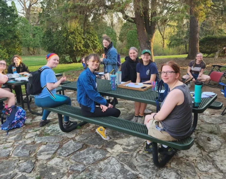 A group of people sit and smile around picnic tables outdoors in a wooded park area near Loke Hall.