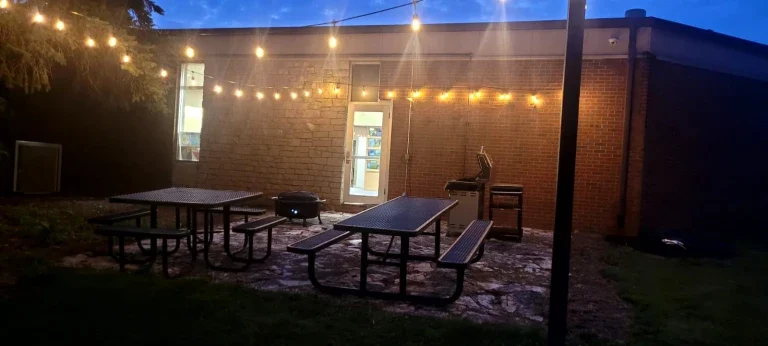 Outdoor patio with picnic tables, string lights, and the iconic brick facade of Loke Hall in the background at dusk.