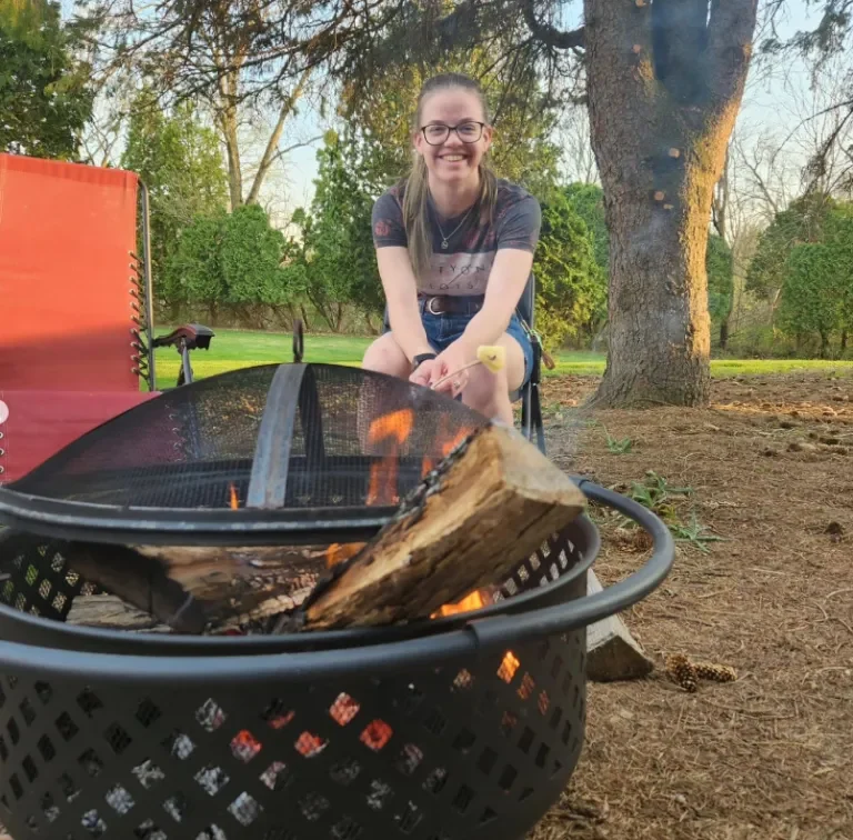Woman smiling and roasting a marshmallow over a fire pit outdoors, with the scenic trees near Loke Hall in the background.