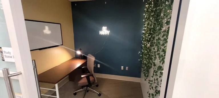 A cozy study nook in Loke Hall with a desk, chair, lamp, "Be Happy" sign, and string lights on a wall draped with hanging vines.