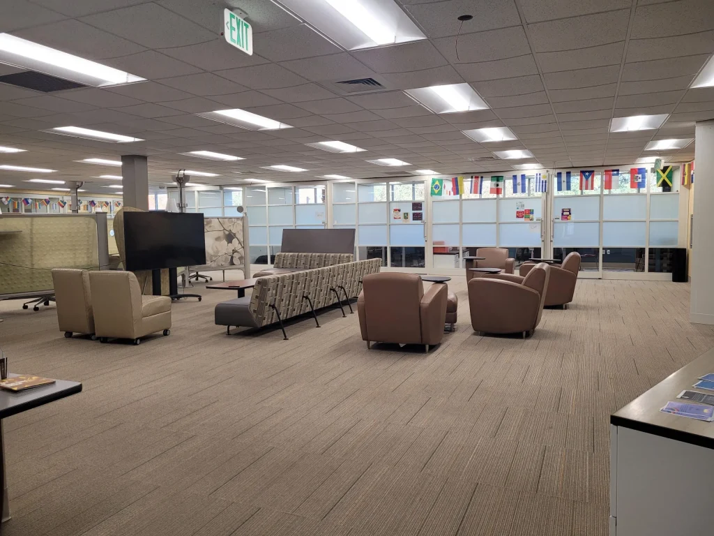 Spacious lounge in Loke Hall with sofas, chairs, world flags, and a large TV in a brightly lit room with carpeted floors.