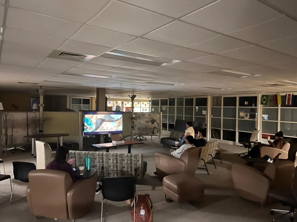 People sit on couches and chairs in the dimly lit lounge of Loke Hall, watching a TV screen at the front of the room.