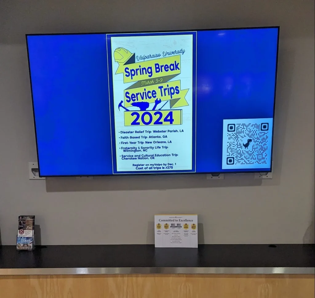 A TV in Harre Union displays a flyer for Valparaiso University’s 2024 Spring Break Service Trips and a QR code.