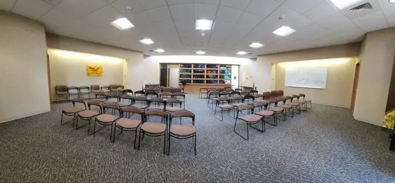 Loke Hall’s conference room features rows of brown chairs facing a bookshelf and whiteboard under bright ceiling lights.
