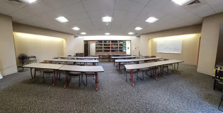 Classroom in Loke Hall with several tables and chairs arranged in rows, a whiteboard on the wall, and shelves at the back.