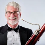 Smiling older man, Eric Kuehner, in a tuxedo holds a bassoon against a plain light background.