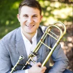Corey Sansolo, smiling in a gray blazer, holds a trombone outdoors with a blurred green background.