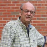 Ronald Janke, Ph.D., an older man in glasses and a plaid shirt, stands in front of a brick wall, looking at the camera.