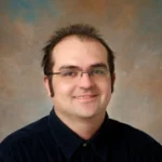 Daniel Maxin, a man with glasses and short dark hair, smiles at the camera against a brown and blue mottled background.