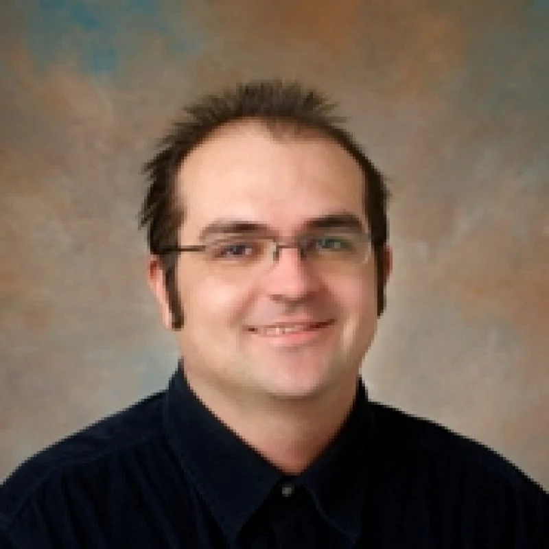 Daniel Maxin, a man with glasses and short dark hair, smiles at the camera against a brown and blue mottled background.
