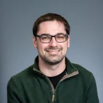 Paul F. Smith, a man with short brown hair and glasses, smiles while wearing a green pullover in front of a plain gray background.