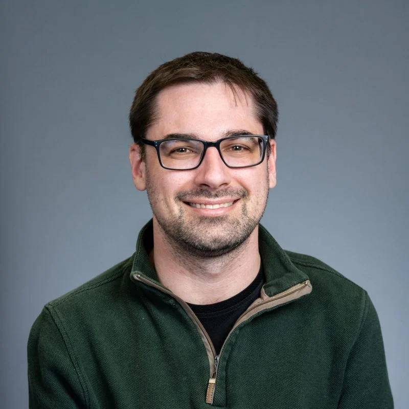 Paul F. Smith, a man with short brown hair and glasses, smiles while wearing a green pullover in front of a plain gray background.